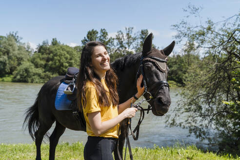 Glücklicher Trainer mit schwarzem Pferd in der Nähe des Sees an einem sonnigen Tag stehend - NDEF01649