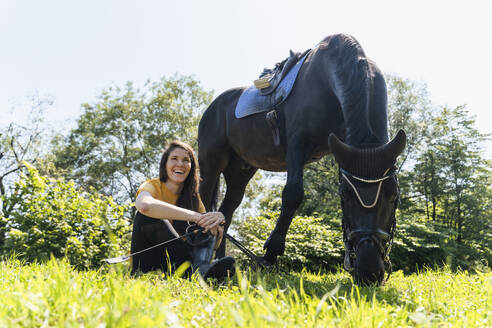 Glückliche Lehrerin sitzt bei einem grasenden schwarzen Pferd an einem sonnigen Tag - NDEF01650