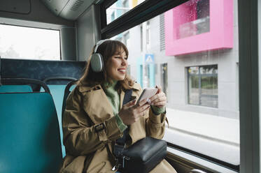 Smiling woman with wireless headphones and smart phone looking out through bus window - ALKF01119