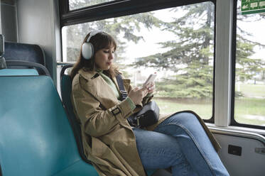 Woman listening to music through wireless headphones using smart phone in bus - ALKF01120