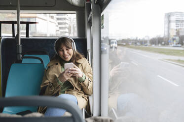 Happy woman texting through mobile phone listening to music in bus - ALKF01121