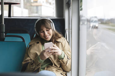 Lächelnde Frau, die eine SMS über ihr Mobiltelefon schreibt und im Bus Musik hört - ALKF01122