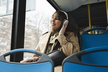 Lächelnder junger Fahrgast, der im Bus sitzend über drahtlose Kopfhörer Musik hört - ALKF01135