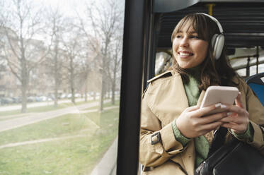 Glücklicher Fahrgast mit drahtlosen Kopfhörern, der durch das Fenster eines Busses nach draußen schaut - ALKF01136