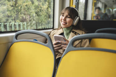 Happy woman wearing wireless headphones looking out through window sitting in bus - ALKF01142