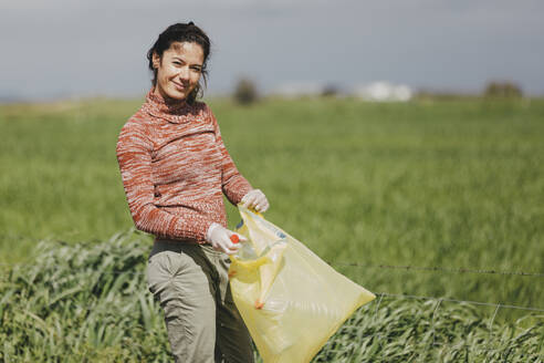 Smiling woman collecting bottles in plastic bag at countryside - DMGF01321