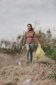 Woman holding plastic bag and collecting bottles at countryside - DMGF01324