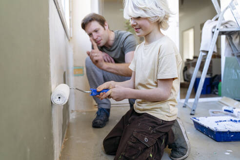 A 6 years old boy with blond long hair is painting a wall with a roller brush and his father a man with European appearance is looking at him - NJAF00939