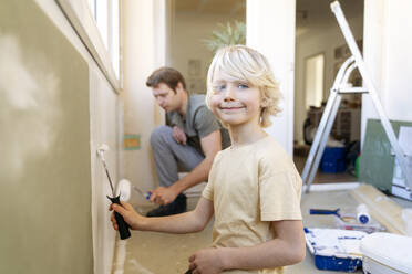 Lächelnder Junge, der mit einem Roller in der Nähe seines Vaters zu Hause eine Wand bemalt - NJAF00945