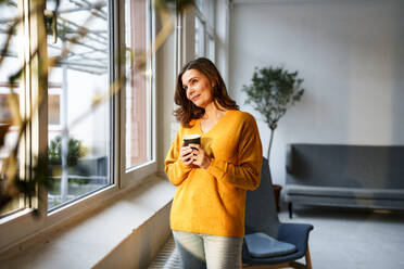 Mature woman holding coffee cup looking out through window at home - KNSF10215