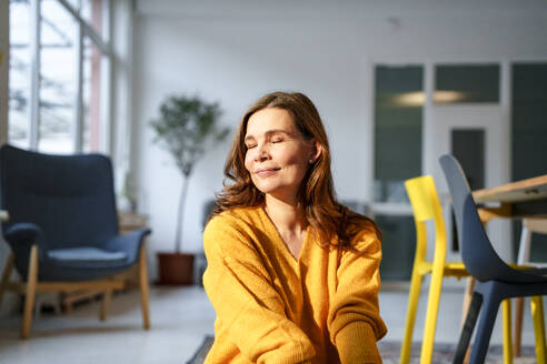 Lächelnde reife Frau mit geschlossenen Augen sitzt unter Sonnenlicht im Wohnzimmer zu Hause - KNSF10222
