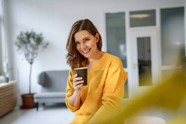 Glückliche reife Frau sitzt mit Kaffeetasse im Wohnzimmer zu Hause - KNSF10228