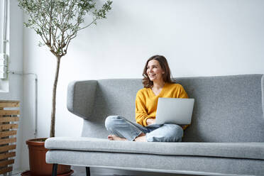 Glückliche Frau sitzt im Schneidersitz auf dem Sofa mit Laptop im Wohnzimmer zu Hause - KNSF10230