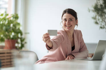 Glückliche Geschäftsfrau sitzt mit Laptop am Tisch und zeigt ihre Visitenkarte bei der Arbeit zu Hause - KNSF10260