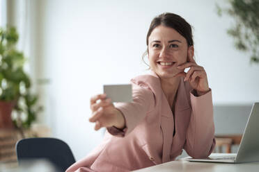 Lächelnde Geschäftsfrau, die mit einem Laptop an einem Tisch sitzt und eine Visitenkarte zeigt und zu Hause arbeitet - KNSF10261