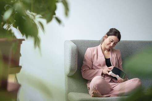 Lächelnde Geschäftsfrau, die mit einem Virtual-Reality-Simulator auf dem Sofa im Heimbüro sitzt - KNSF10268