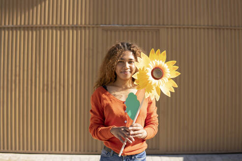 Lächelnde Frau mit künstlicher Sonnenblume vor einer Metallwand - MGRF01118