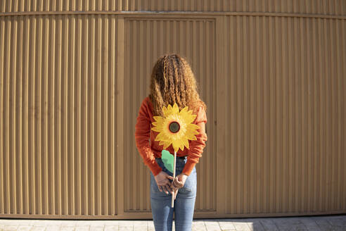 Junge Frau hält künstliche Sonnenblume vor einer Metallwand - MGRF01120