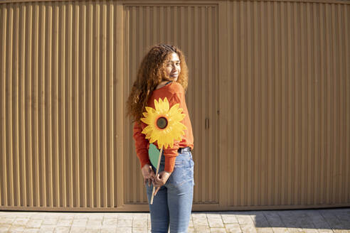 Junge Frau hält künstliche Sonnenblume hinter dem Rücken vor einer Metallwand - MGRF01121