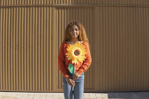 Lächelnde Frau mit künstlicher Sonnenblume vor einer Metallwand an einem sonnigen Tag - MGRF01124