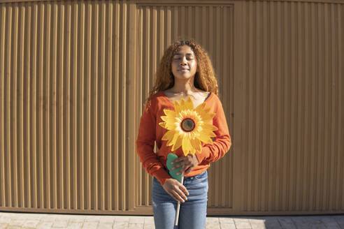 Junge Frau mit geschlossenen Augen, die eine künstliche Sonnenblume hält und vor einer Metallwand steht - MGRF01126