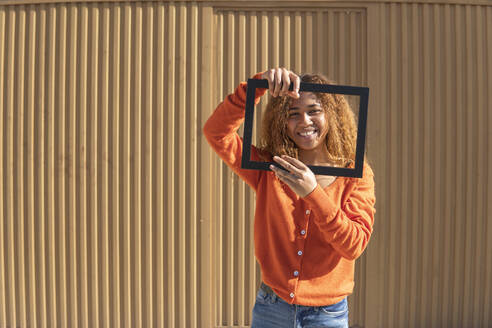 Glückliche Frau mit Bilderrahmen vor einer Metallwand stehend - MGRF01128