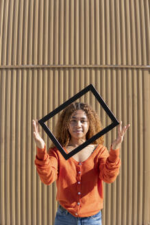 Junge Frau mit Bilderrahmen vor einer Metallwand stehend - MGRF01129