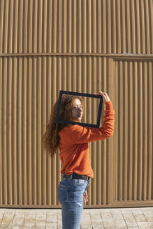 Junge Frau mit Bilderrahmen auf der Schulter vor einer Metallwand stehend - MGRF01132