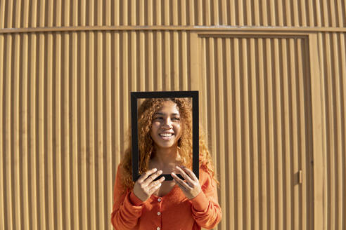 Lächelnde junge Frau, die durch einen Bilderrahmen wegschaut und vor einer Metallwand steht - MGRF01134