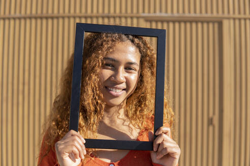 Lächelnde junge Frau mit lockigem Haar schaut durch einen Bilderrahmen - MGRF01135