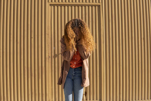 Junge Frau, die ihr Gesicht mit den Händen bedeckt, vor einer Metallwand - MGRF01138