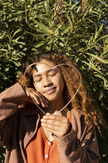 Curly-haired black girl posing in a brown jacket and a metal heart. Natural leaves background. Granada, Spain. - MGRF01144