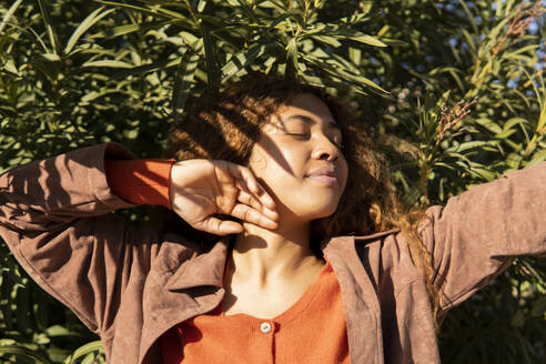 Curly-haired black girl posing relaxed and enjoying, wearing a brown jacket. Natural leaves background. Granada, Spain. - MGRF01146