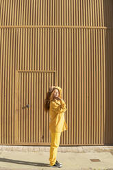 Junge Frau in gelber Freizeitkleidung steht auf dem Fußweg vor einer Metallwand - MGRF01156