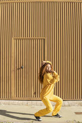 Frau in gelber Freizeitkleidung tanzt vor einer Metallwand an einem sonnigen Tag - MGRF01157