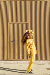 Glückliche Frau mit lockigem Haar und gelber Freizeitkleidung tanzt vor einer Metallwand - MGRF01158