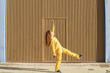 Junge Frau in gelber Freizeitkleidung tanzt auf dem Fußweg vor einer Metallwand - MGRF01159