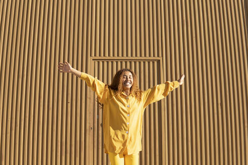 Glückliche, lockige Frau in gelber Freizeitkleidung steht mit ausgestreckten Armen vor einer Metallwand - MGRF01160