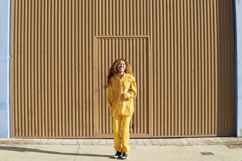 Glückliche Frau in gelber Freizeitkleidung steht vor einer Metallwand an einem sonnigen Tag - MGRF01162
