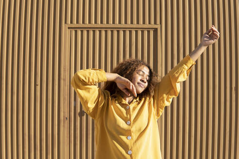 Frau in gelber Freizeitkleidung tanzt an einem sonnigen Tag vor einer Mauer - MGRF01164