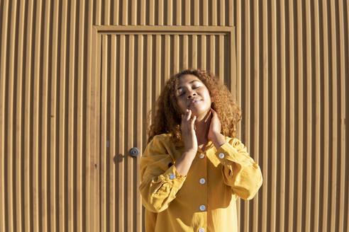 Frau in gelber Freizeitkleidung tanzt vor einer orangefarbenen Wand an einem sonnigen Tag - MGRF01165
