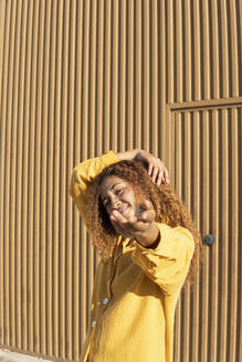 Lächelnde Frau in gelber Freizeitkleidung tanzt vor einer orangefarbenen Wand - MGRF01166