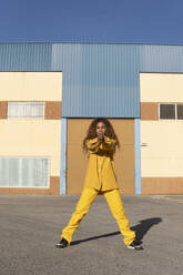 Junge Frau in gelber Freizeitkleidung steht breitbeinig auf einem Fußweg - MGRF01174