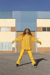 Junge Frau in gelber Freizeitkleidung steht mit ausgestreckten Armen auf einem Fußweg - MGRF01175