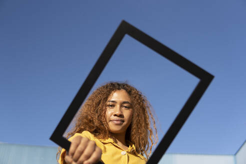 Curly-haired black girl dressed in yellow pants and shirt, posing with a photo frame. Granada, Spain. - MGRF01187