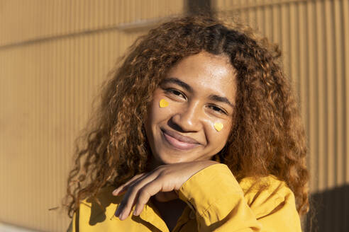 Black girl with curly hair dressed in yellow pants and shirt, posing with yellow hearts stuck on her cheeks. Granada, Spain. - MGRF01191