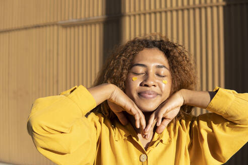 Black girl with curly hair dressed in yellow pants and shirt, posing with yellow hearts stuck on her cheeks. Granada, Spain. - MGRF01192