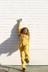 Junge Frau in gelber Freizeitkleidung lehnt sich mit erhobener Hand an einen weißen Fensterladen - MGRF01201