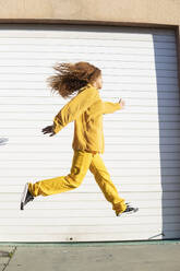 Aufgeregte junge Frau in gelber Freizeitkleidung springt vor einem weißen Fensterladen - MGRF01208