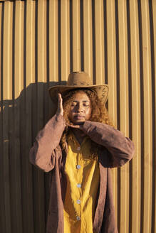 Frau mit geschlossenen Augen, die eine Jacke und einen Cowboyhut trägt und vor einer orangefarbenen Metallwand steht - MGRF01238